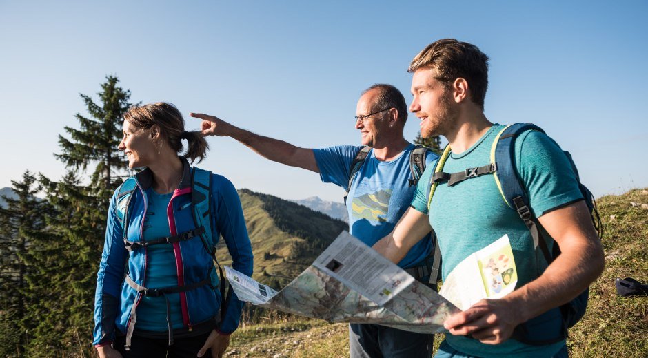 Wanderer steigen einen Bergpfad hinauf, umgeben von malerischen Alpenlandschaften und grünen Wiesen. Im Hintergrund sind sanfte Berggipfel und Wolken zu sehen. © Tourismus Hörnerdörfer, F. Kjer
